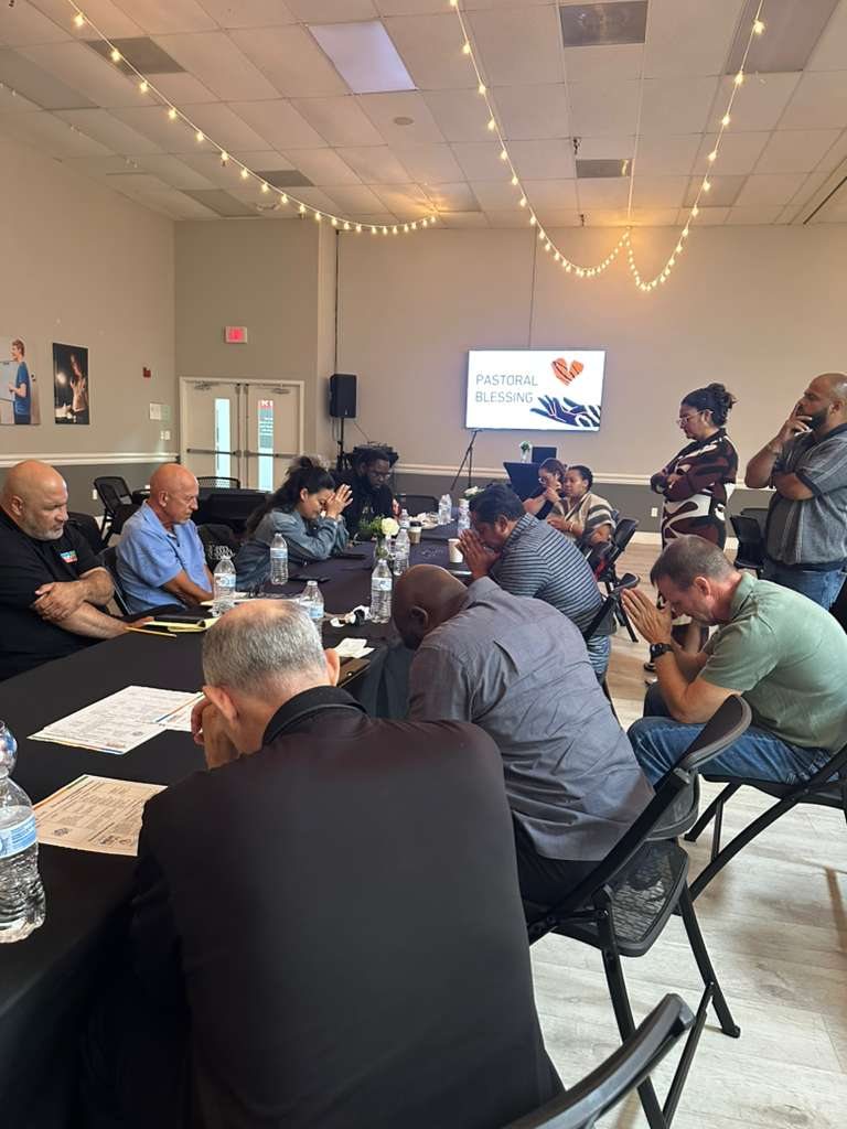 Haitian, Latino, and Black pastors and ministry leaders praying together around a table at a pastoral gathering